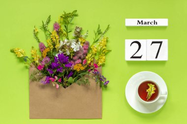 Ahşap küpler takvim 27 Mart. Otlar çay Fincan, yeşil arka plan üzerinde çok renkli çiçekler ile zanaat zarf.
