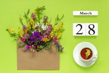 Ahşap küpler takvim 28 Mart. Otlar çay Fincan, yeşil arka plan üzerinde çok renkli çiçekler ile zanaat zarf.