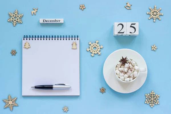 木製カレンダー 12月25日 マシュマロとスターアニスのココアカップ 青い背景にペンと雪片と空のオープンメモ帳 — ストック写真