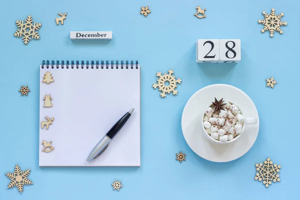 木製カレンダー 12月28日 マシュマロとスターアニスのココアカップ 青い背景にペンと雪片と空のオープンメモ帳 — ストック写真