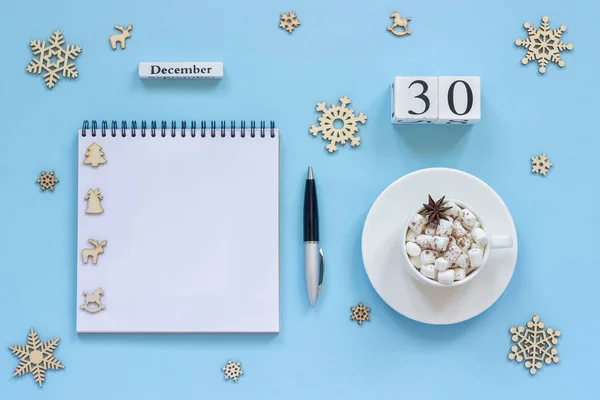 12月30日の白い木製のカレンダーキューブ マシュマロとスターアニスのココアカップ 青い背景にペンと雪片と空のオープンメモ帳 — ストック写真