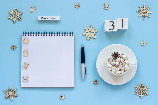12月31日の白い木製のカレンダーキューブ マシュマロとスターアニスのココアカップ 青い背景にペンと雪片と空のオープンメモ帳 — ストック写真