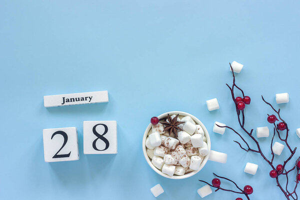 White wooden calendar cubes with January 28. Cup of cocoa, marshmallows and decorative branch with red berries on blue background 