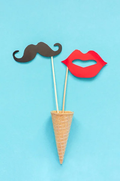Couple of paper mustache and lips props on stick in ice cream waffle cone on blue background