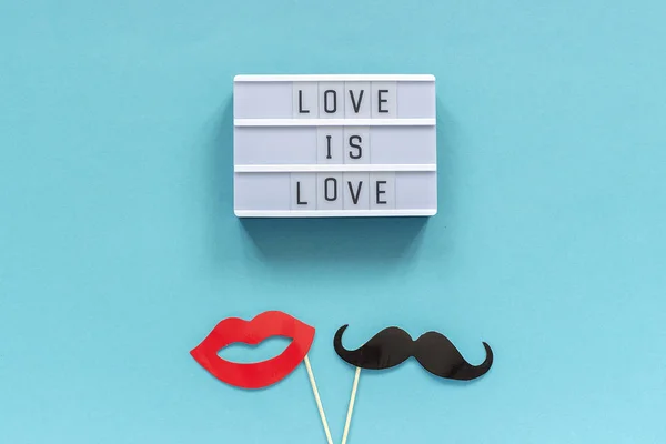 Light box with text Love is love and paper mustache and lips props on on blue background