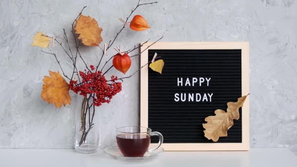 Χαρούμενο Κυριακάτικο κείμενο σε μαύρο γράμμα πίνακα και μπουκέτο από κλαδιά με κίτρινα φύλλα σε μανταλάκια σε βάζο και φλιτζάνι τσάι στο τραπέζι πρότυπο για καρτ ποστάλ, ευχετήρια κάρτα concept Hello φθινόπωρο Κυριακή — Αρχείο Βίντεο