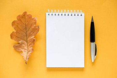 Sonbahar kompozisyonu. Beyaz boş not defteri, kurutulmuş portakal meşe yaprağı ve sarı arka plan üzerinde kalem. Üst görünüm Düz lay Mockup Şablon u tasarımınız için