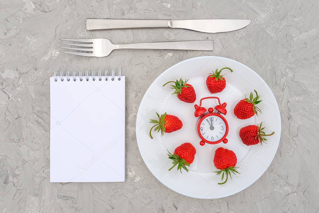 Red ripe strawberries berry on white plate, cutlery, red alarm clock and blank notepad on gray stone background table. Top view, flat lay, Mockup. Concept diet and detox time or summer menu time