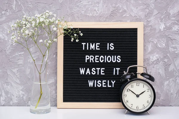 Time is precious waste it wisely. Motivational quote on letterboard, black alarm clock, flower in vase on table against stone wall. Concept quote of the day — Stock Photo, Image