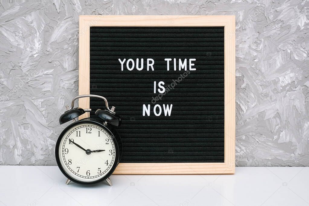 Your time is now. Motivational quote on letter board and black alarm clock on table against stone wall. Concept inspirational quote of the day