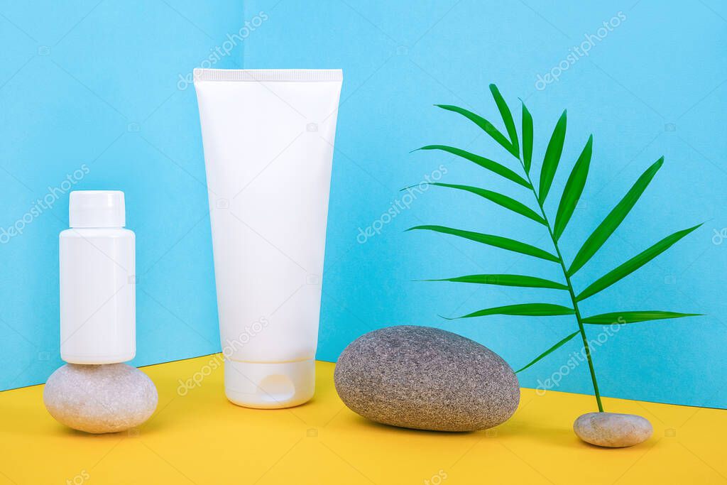 White blank cosmetics bottles, tube of cream, lotion for body, face, hand or other cosmetic product and gray rocks, palm leaf on blue yellow background. Mockup Front view, Corner composition .