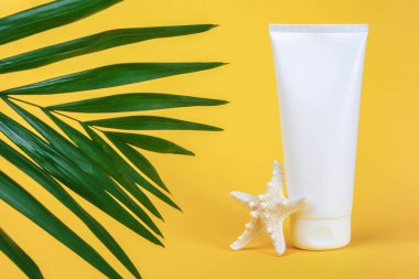 Güneş kremi sürmüş beyaz boş kozmetik tüp, yüz veya vücut için güneş kremi, denizyıldızı ve sarı arka planda yeşil brunch palmiyesi. Yaz tatilinde cilt bakımını kavramsallaştır Ön görünüm kopyalama alanı.