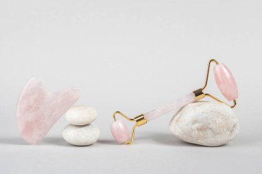 Kristal gül kuvars yüz silindiri ve masaj aleti taşların üzerinde yeşil Gua sha, gri arka plan. Evde doğal kaldırma ve tonlama tedavisi için yüz masajı. Güzellik konsepti Ön görünüm.