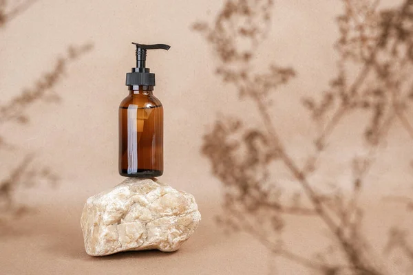 Brown glass bottle with pump of cosmetic products on stone framed by dried plant flowers on beige craft paper background. Natural Organic Spa Cosmetic Beauty concept Front view Mock up.