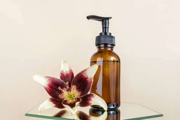Brown glass bottle with pump of cosmetic products and lily flower on mirror, beige background. Natural Organic Spa Beauty Cosmetic concept Mockup.
