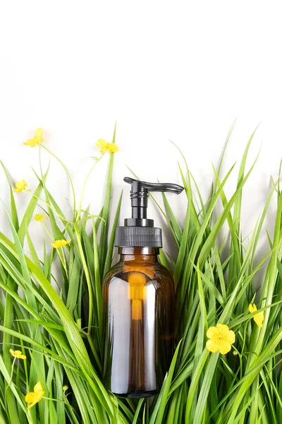 Brown glass bottle with pump of cosmetic products among the green grass, yellow flowers on white background. Natural Organic Spa Beauty Cosmetic concept Mockup.