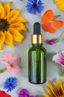 Yaşlanma önleyici kolajen, yüz serumu ya da gri arka plandaki renkli çiçekler arasında yeşil cam şişedeki diğer kozmetik ürünler. Doğal Organik Spa Kozmetik Konsepti Üst Görünüm.