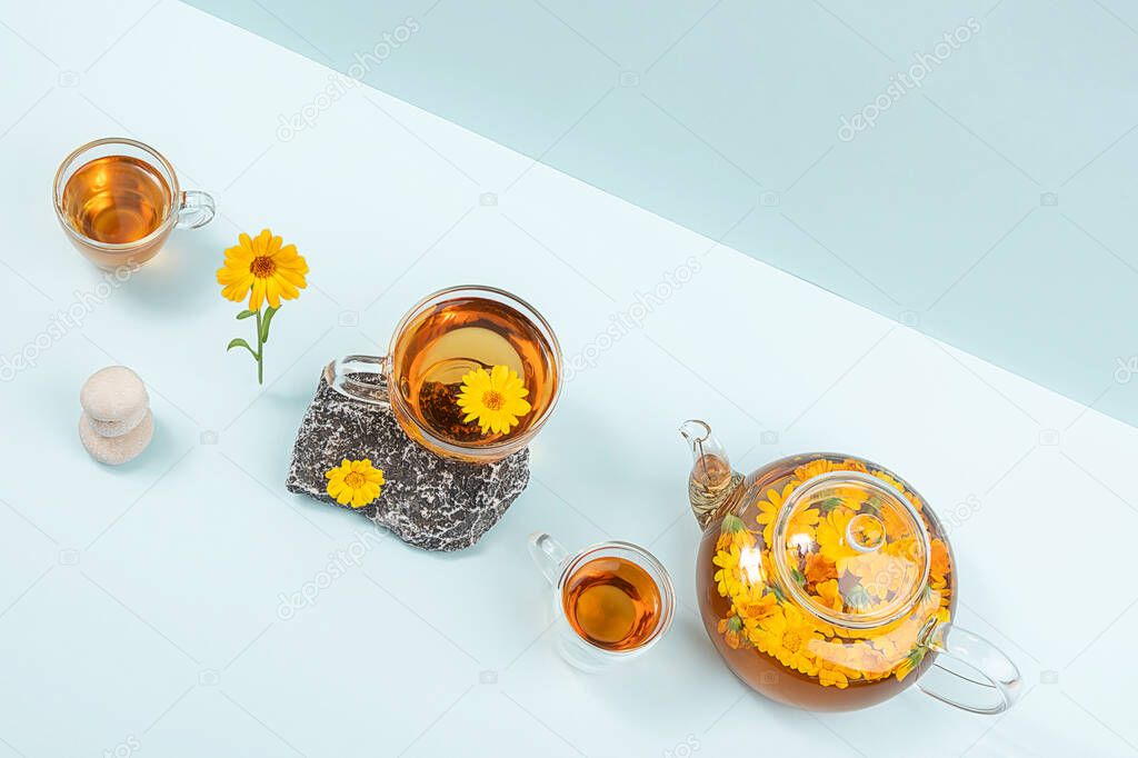 Cup of herbal tea and transparent teapot with calendula flowers on blue background. Calendula Tea Benefits Your Health concept. Top view Flat lay.