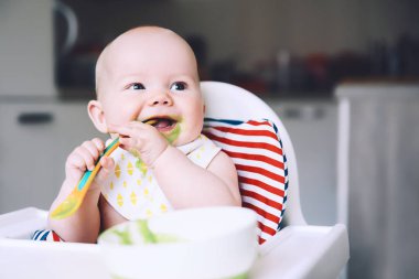 Besleme. Dağınık gülümseyen bebek yüksek sandalye bir kaşıkla yemek. Bebeğin ilk katı yiyecek. Anne küçük çocuk püre kaşıkla besleme. Günlük rutin. Parmak gıda. Sağlıklı çocuk beslenme.