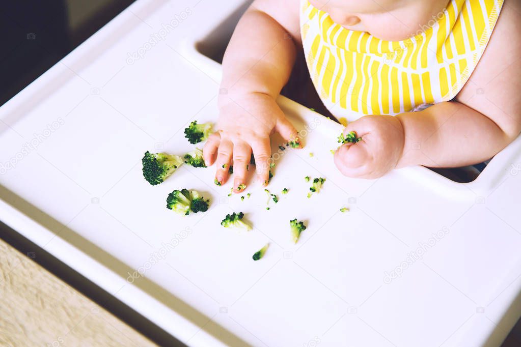 Baby's first solid food. Messy smiling baby eats and tastes with fingers vegetables broccoli in high chair. Healthy child nutrition. Mother gives to try foods to feed little child 7 months old