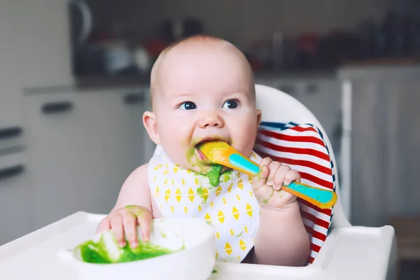 乱雑な微笑の赤ん坊は高い椅子でスプーンで食べる 赤ちゃんの最初の固形食 母のピューレのスプーンで小さな子供を供給します 毎日のルーチン フィンガー フード 健康な子供の栄養 — ストック写真