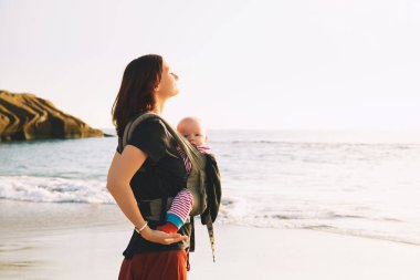 Bebek ve anne yaz gün denizde üzerinde. Mutlu bir aile doğa açık havada yürüyüş. Çocuk bir taşıyıcı sırt çantası içinde. Kadın ve bebeğinin adanın Tenerife, İspanya'nın kıyı şeridi okyanus üzerinde. Avrupa'yı gezersin