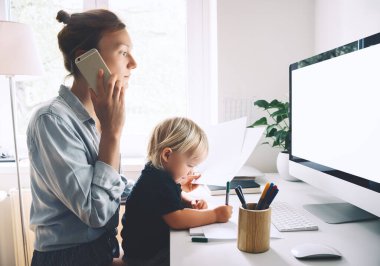 Modern anneler, hastalık izninde iş ve çocuk eğitimi arasında denge kurar. Annem evde çocuğuyla çalışıyor. Telefonlara cevap veren bir kadın kucağında bir bebekle bilgisayarla çalışıyor..
