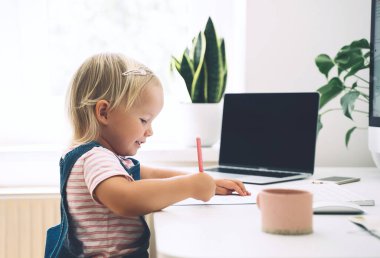 Küçük kız ders çalışıyor ve masada resim çiziyor. Anaokulu çocuğu erken gelişimde bilgisayar kullanıyor ya da evde eğlence yapıyor. Çocuklar evde eğitim ve uzak online eğitim. Çocuklar çevrimiçi öğrenme.