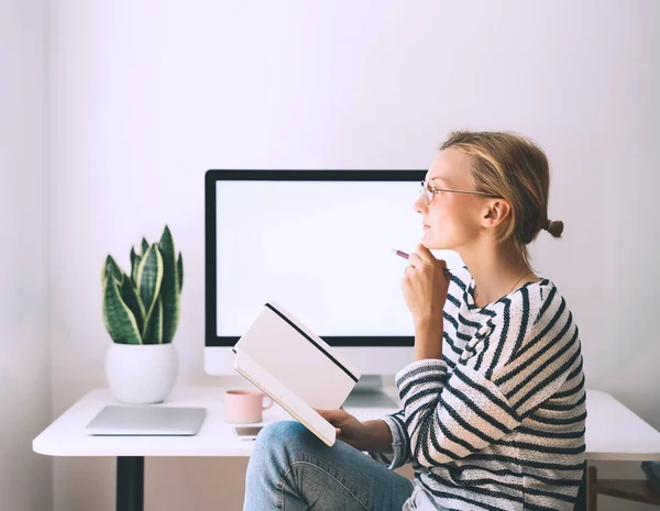 Young woman at home office. Girl wearing in glasses works with new startup project. Female thinking and writing in planner or diary at workplace of minimalist style with computer.