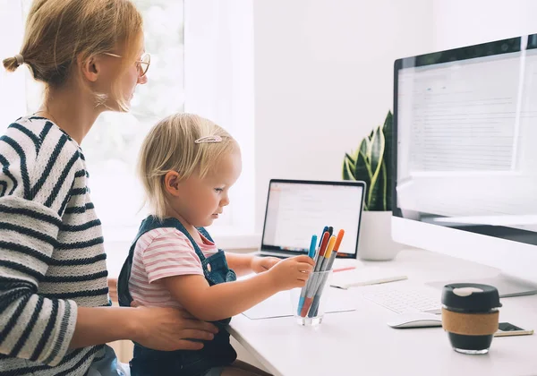 Modern mom balances between work and child home schooling on sick leave or quarantine. Mother works from home with kid. Woman responding on phone calls, working on computer with a toddler on her lap.