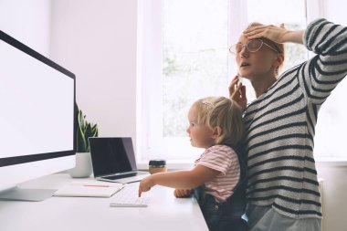 Modern anneler, hastalık izninde iş ve çocuk eğitimi arasında denge kurar. Annem evde çocuğuyla çalışıyor. Telefonlara cevap veren bir kadın kucağında bir bebekle bilgisayarla çalışıyor..