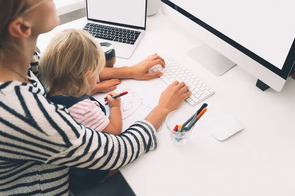 Modern mom balances between work and child home schooling on sick leave or quarantine. Mother works from home with kid. Woman responding on phone calls, working on computer with a toddler on her lap.