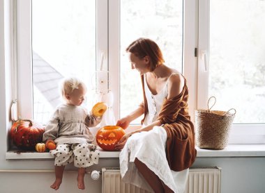 Çocuk sahibi minimalist bir aile Cadılar Bayramı için hazırlanıyor. Mutlu anne ve küçük kızı pencerenin kenarında oturmuş balkabağı kafası oyuyorlar Jack feneri ve cadılar bayramı süslemesi yapıyorlar..