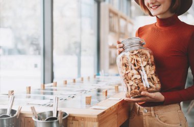 Plastiksiz marketin arka planında büyük cam bir kavanoz taşıyan bir kadın. Plastik ambalajsız yerel yiyecekler alan bir kadın. Sıfır atık dükkanı veya yaşam tarzı kavramı.