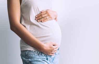 Gündelik giysiler içinde hamile bir kadın beyaz arka planda el ele tutuşuyor. Anne doğumu bekliyor. Kadınlar hamileliğe hazırlanır. Hamilelik kavramı, doğum öncesi dönem, annelik sağlığı.
