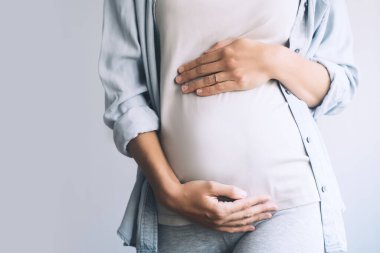 Hamile kadın beyaz arka planda göbeğin üstünde el ele tutuşuyor. Anne doğumu bekliyor. Kadınlar hamileliğe hazırlanır. Hamilelik kavramı, jinekoloji, doğum öncesi dönem, annelik sağlığı..