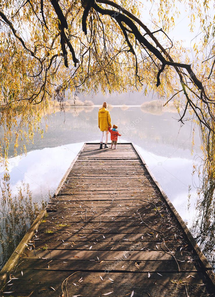 Woman with toddler on wooden pier bridge near by lake surrounded by branches of willow tree in autumn time. Mother and child together in countryside outdoors. Family in nature background.