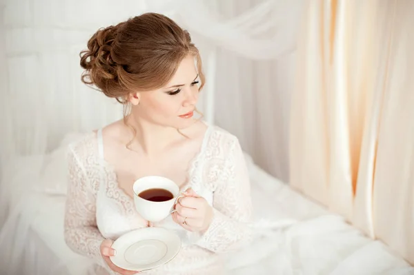 Girl holding a white coffee mug. In a white coat. Coffee in bed. Morning begins with coffee. Coffee in the hands of the bride