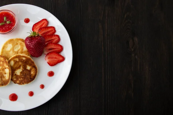 Three pancakes with cheese and strawberries and sauce on a white dish against wood background. Delicious cheese pancake and strawberries for the breakfast or lunch. Tasty meal with berries.