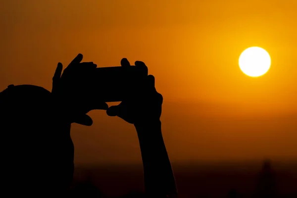 Bir cep telefonu tutan ve gün batımı veya gündoğumu sırasında büyük güneşin fotoğrafçılığı yapma ellerin silueti. Erkek telefonda bir fotoğraf yapar.