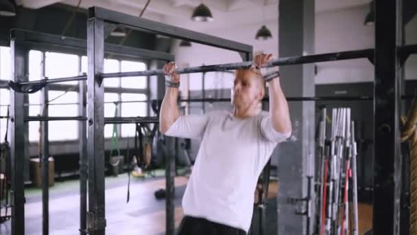 Man pulls up on bar in gym, wears lift and grip gear and white t-shirt — ストック動画