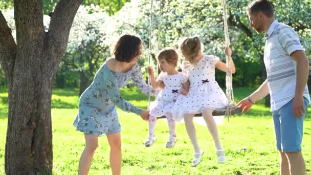 Счастливая молодая семья катает своих девочек на качелях в саду весеннего сезона . — стоковое видео