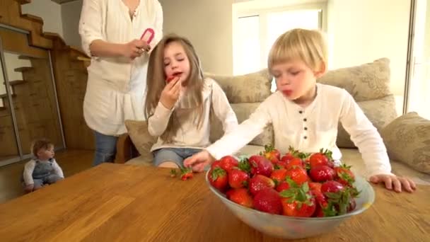 Fille manger fraise tandis que la mère peigne les cheveux et frère. baies mûres rouges dans le plat — Video