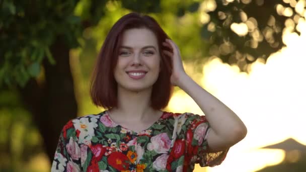 Приваблива дівчина з барвистим квітковим платтям ремонтує волосся і дивиться на камеру — стокове відео