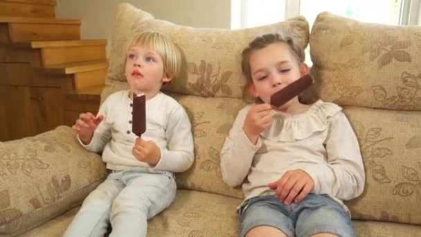 Boy and girl eat ice cream with chocolate on sticks sit on sofa. Handheld shot — Stock Video