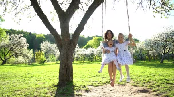 Улыбающаяся мать с двумя дочерьми, качающимися на качелях в прекрасном парке. Статический — стоковое видео