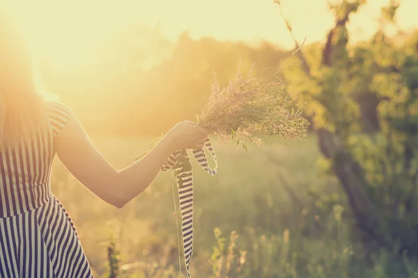 Unrecognizable woman in striped sundress holds bouquet wildflowers in hand, summer concept. Young carefree girl enjoying freedom and sunny nature scene. Picturesque view of countryside at background.