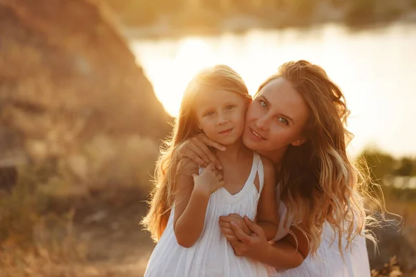 Family, mother and daughter at sunset. The girl bends down and looks at the camera with her daughter. Girls in white dresses. They are blondes. Family time together.