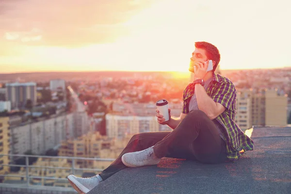 A hipster man drinks coffee and talks on his cell phone. He sits on the roof of a high-rise building at sunset. Portrait in the rays of the setting sun