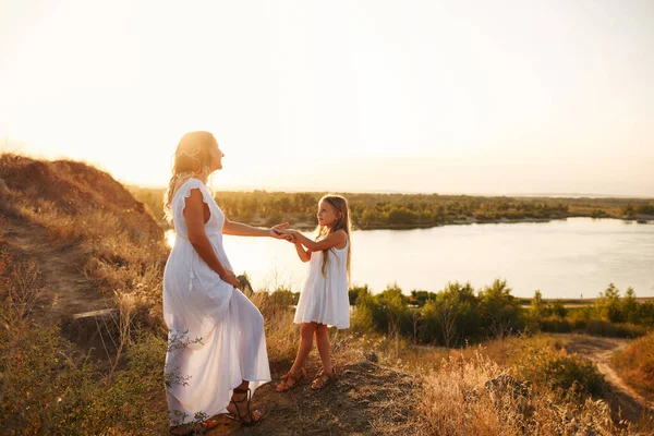 Family, mother and daughter are walking along the river bank. A little girl pulls her mother by the hand. Girls in white dresses. They are blondes. Family time together.